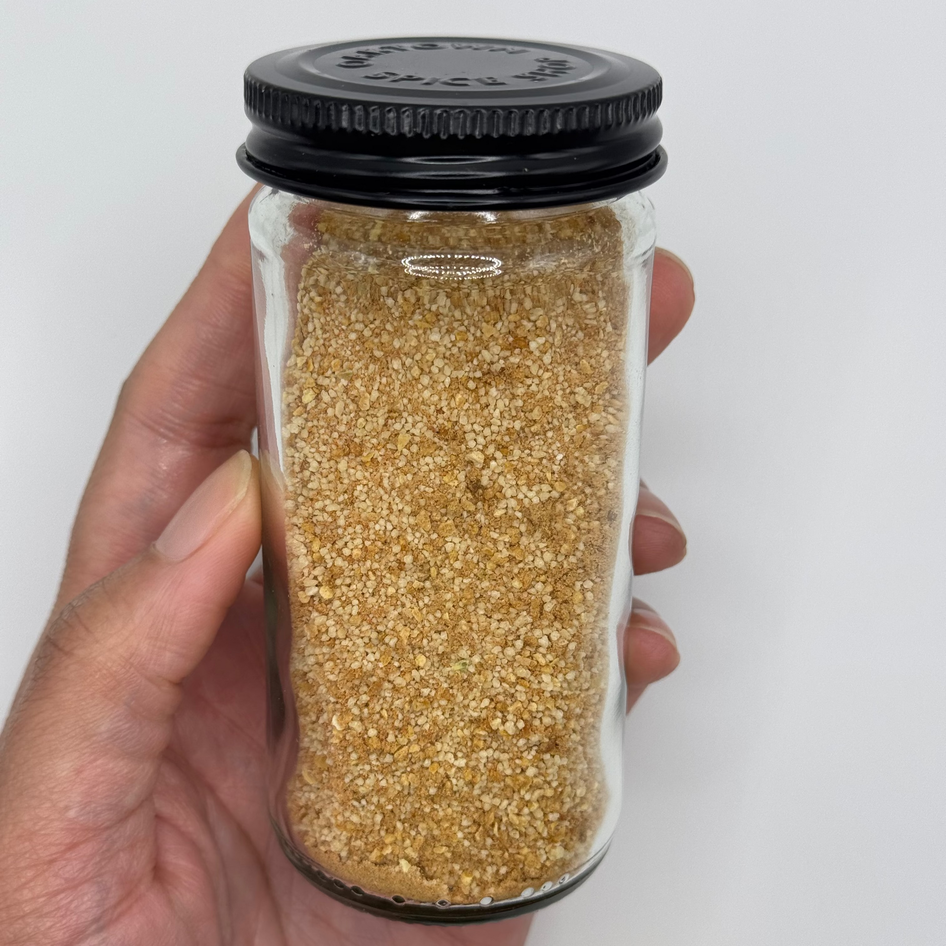Hand holding a glass jar with black lid containing a brown granular substance on a white background