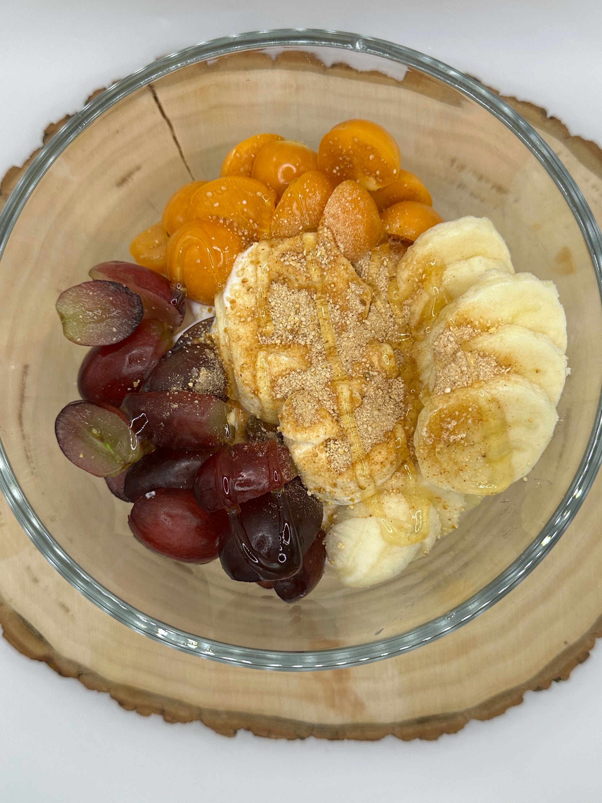 Fruit salad with bananas, grapes, and mandarin oranges in a glass bowl on a wooden coaster.