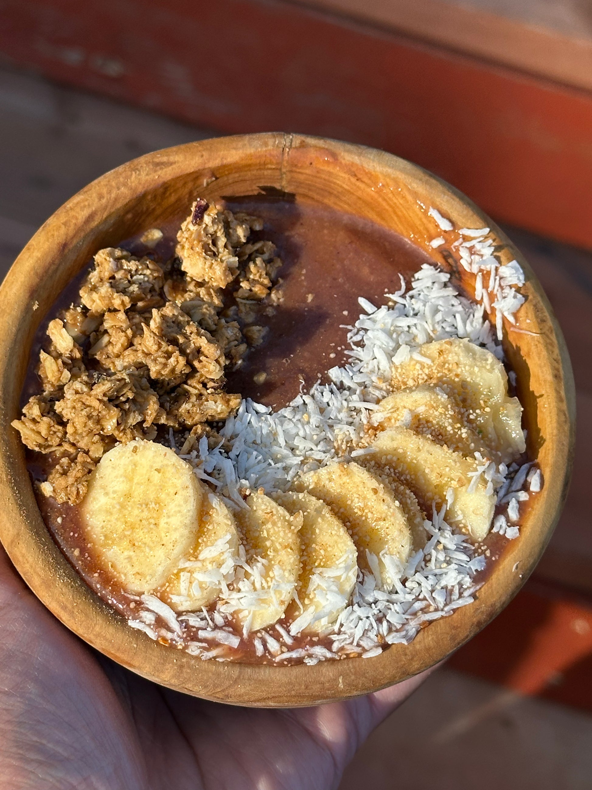 Wooden bowl with a dessert containing chocolate sauce, banana slices, and coconut flakes.