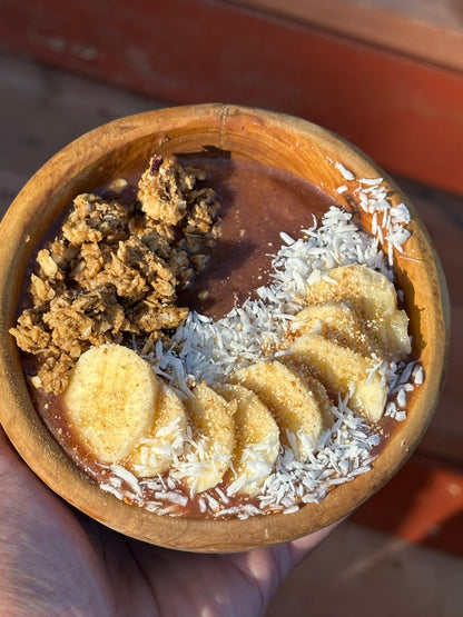 Wooden bowl with a dessert containing chocolate sauce, banana slices, and coconut flakes.