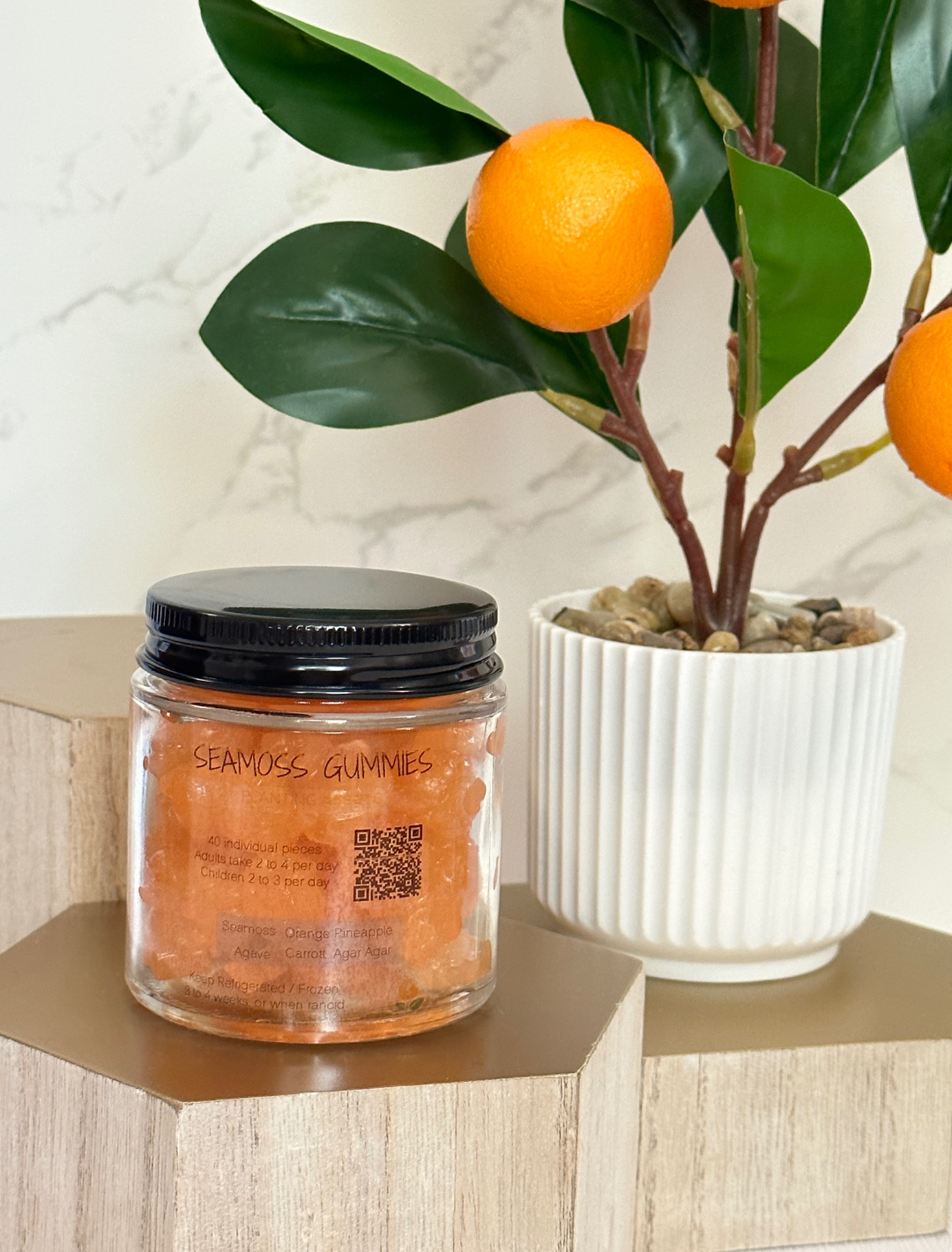 A glass jar containing Seamoss Gummy Supplements, with a label showing the product name and a QR code, placed on a marble surface with an orange plant in the background.
